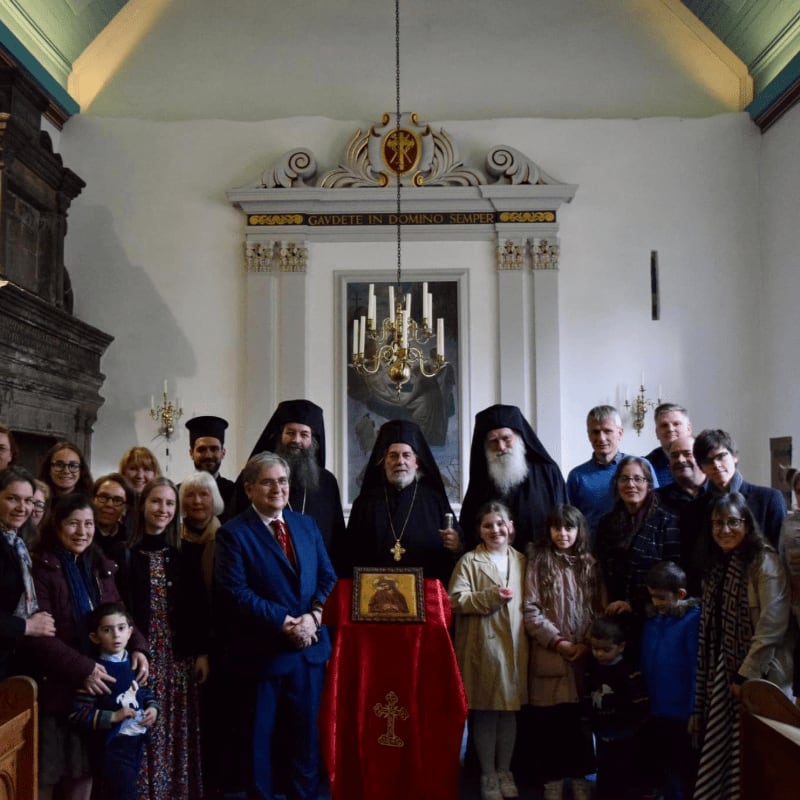 A group of clergy and laypeople stand in front of an altar with an icon, inside a church with ornate architectural details.