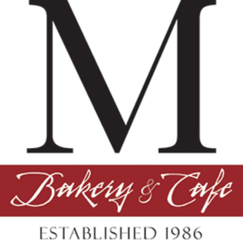 A large black serif "M" is centered above a maroon banner with "Bakery & Cafe" in white script, all set against a white background with "ESTABLISHED 1986" in gray text below the banner.