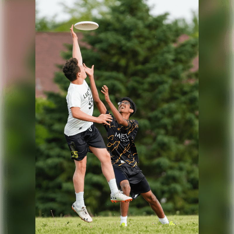 Two young men leap for a frisbee in the foreground, with a blurred background of green trees and a house.