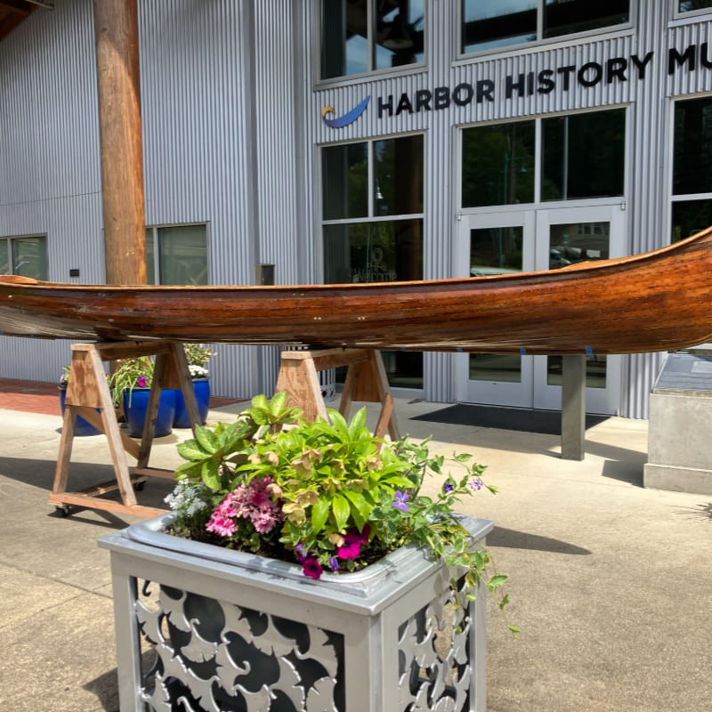 A wooden canoe is displayed on sawhorses in front of a building that reads "HARBOR HISTORY MUSEUM," with a decorative planter in the foreground.
