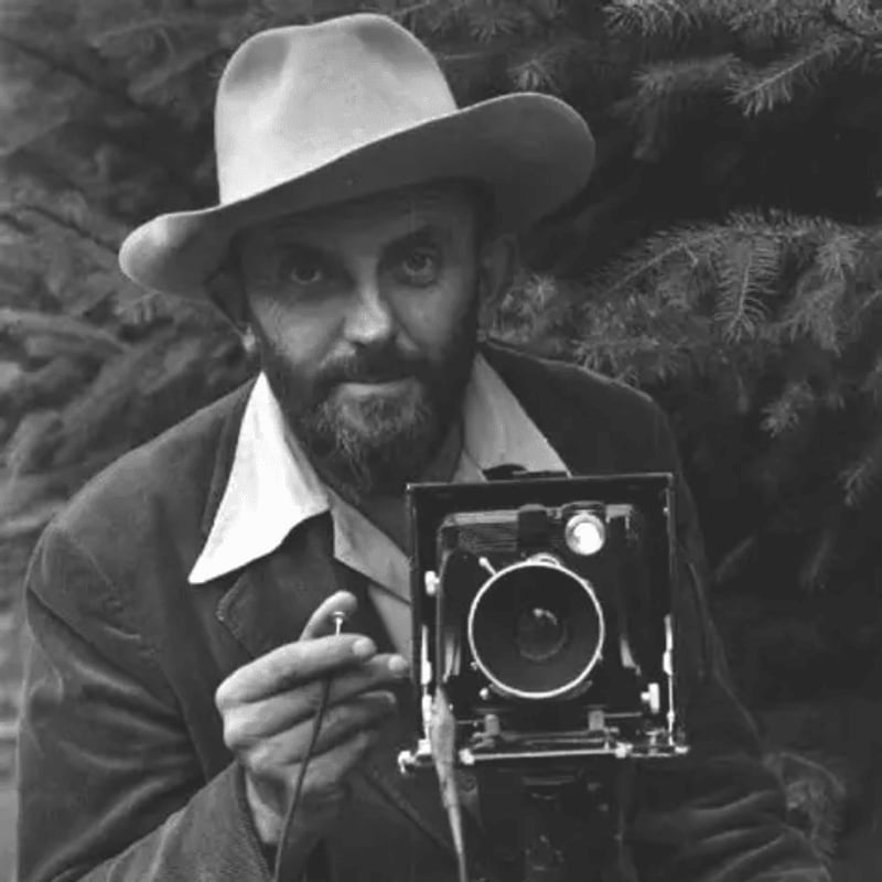 A man in a hat holds a vintage camera in the foreground, with a blurred background of evergreen trees.