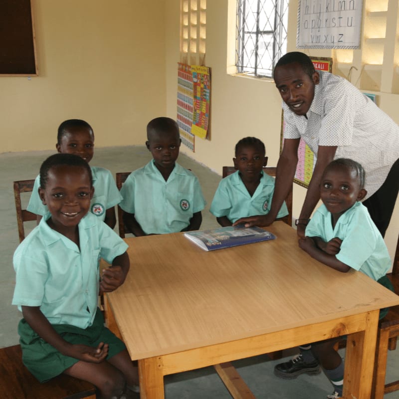 A teacher stands behind a table where five young students in light green uniforms are seated, with colorful educational charts on the wall in the background.