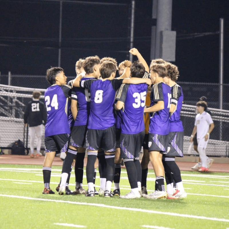 A group of young soccer players in purple jerseys huddle together on a green field with stadium lights and bleachers in the background.