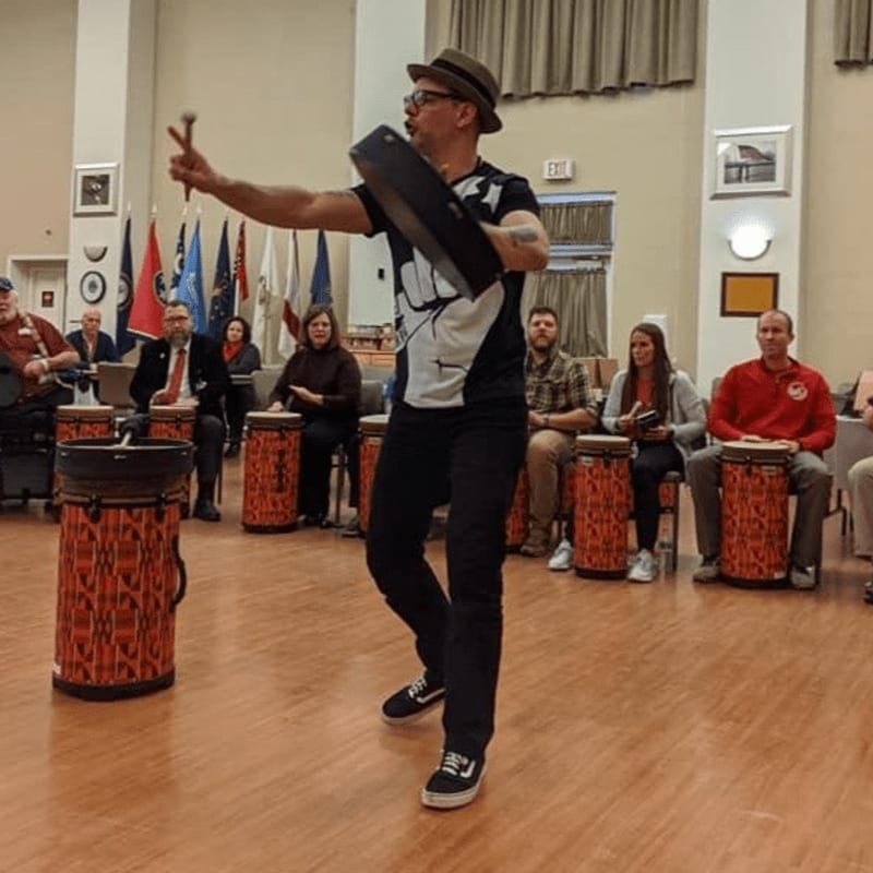 A man in a hat leads a drum circle with colorful drums in the foreground, while an audience watches in the background.