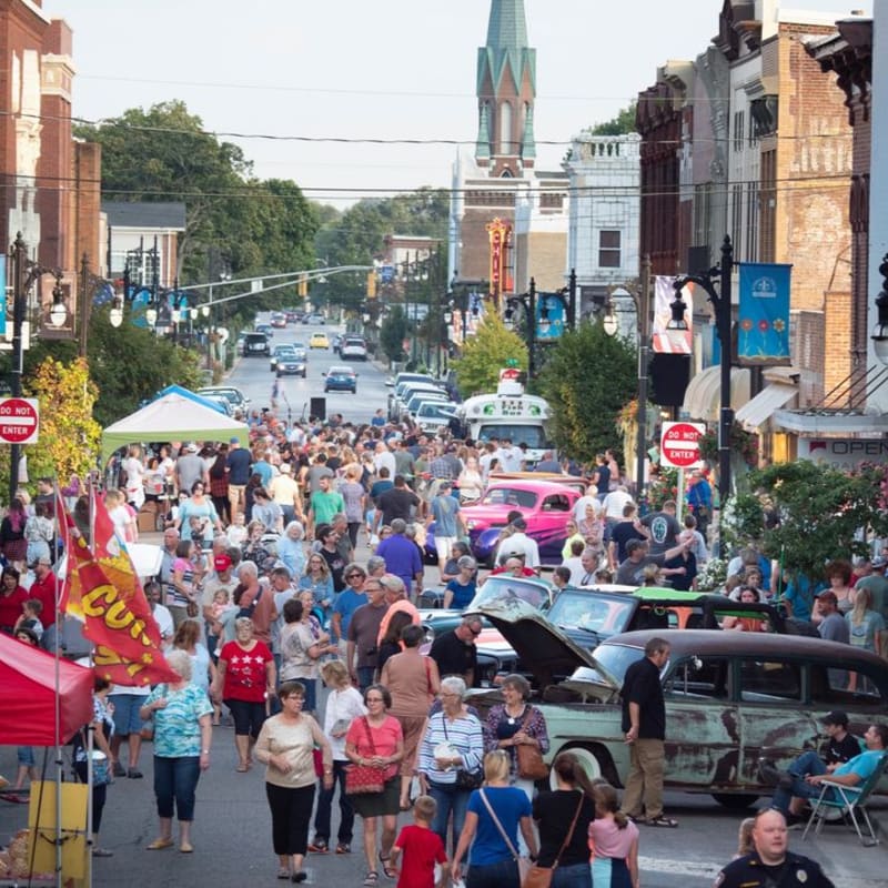 A lively street fair fills the foreground with a crowd of people browsing stalls and vintage cars, while the background shows a quaint town street lined with buildings and a distant church steeple.