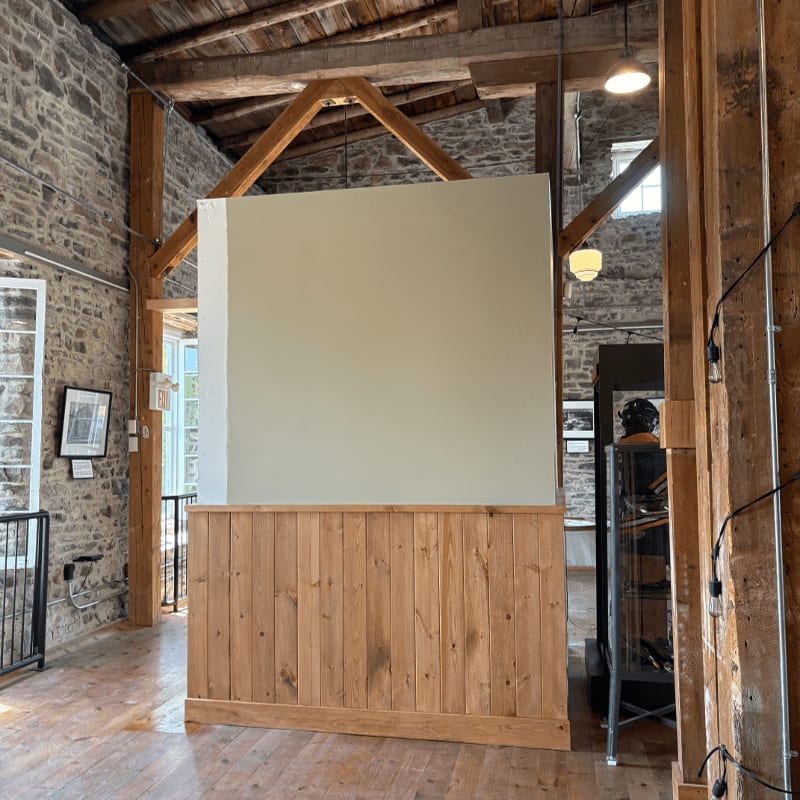 A large, light-colored partition with a wooden base stands in the foreground, while the background reveals a rustic interior with stone walls, exposed wooden beams, and windows.