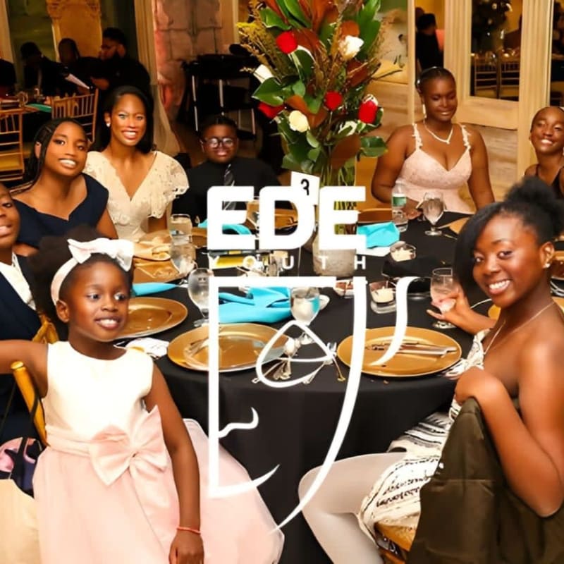A group of smiling people are seated at a formal dinner table in the foreground, with a blurred background of a decorated event space.