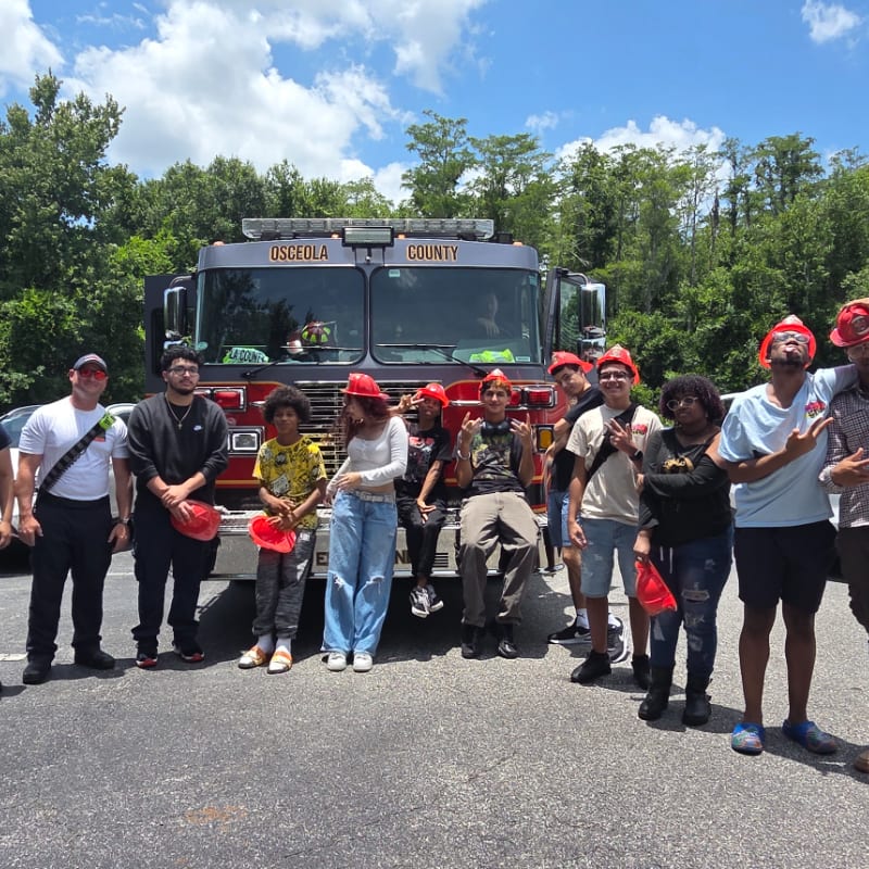 A group of young people in red hard hats stand in front of a fire truck with trees in the background.