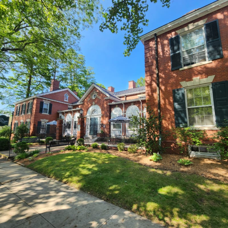A sunlit sidewalk leads past a manicured lawn and a red brick building with large windows and black shutters, backed by lush green trees.
