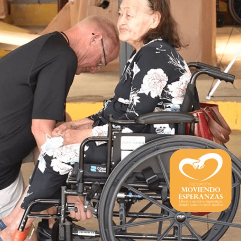A man in a black shirt adjusts the footrest of a woman in a wheelchair, with a logo in the lower right corner and a blurred background of an indoor setting.