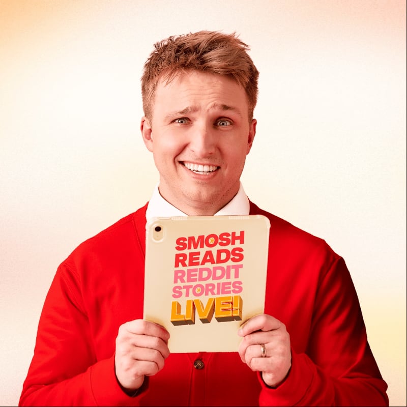 A man in a red cardigan holds a tablet displaying "SMOSH READS REDDIT STORIES LIVE!" against a softly lit, gradient background.
