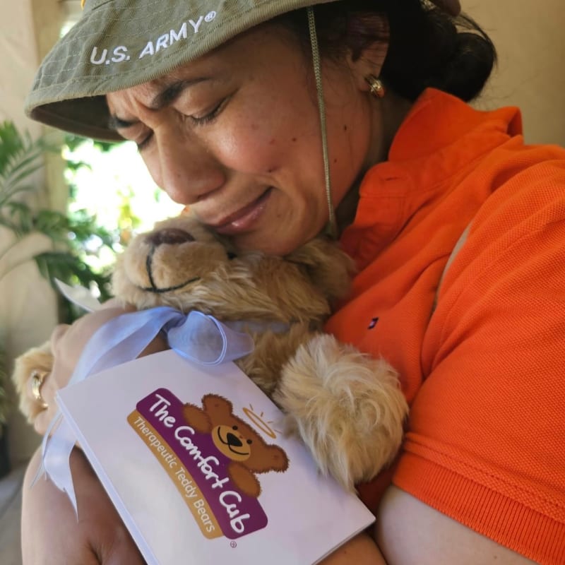 A woman wearing a U.S. Army hat and an orange shirt tenderly hugs a teddy bear with a "The Comfort Cub" tag in front of a blurred green background.