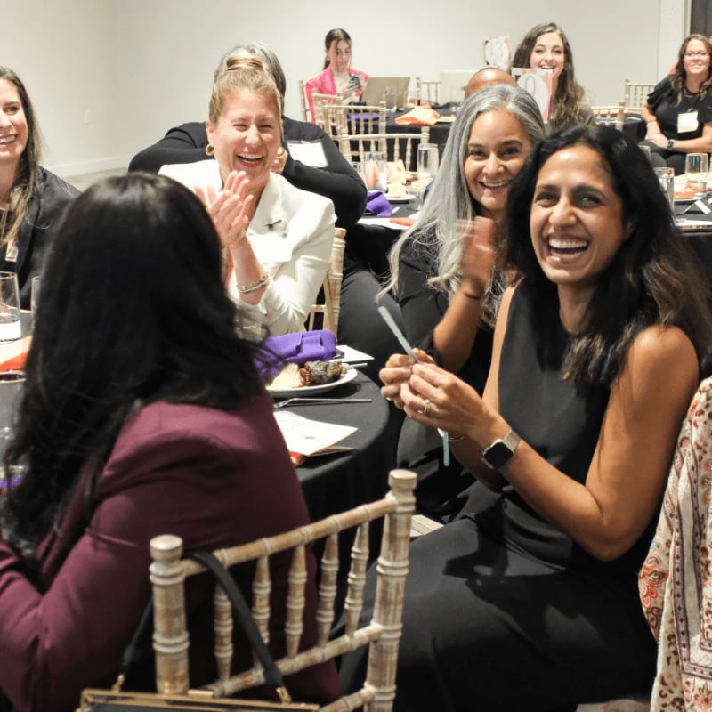 In the foreground, women in formal attire laugh and clap at a table, while in the background, other attendees are seated at tables in a brightly lit room.