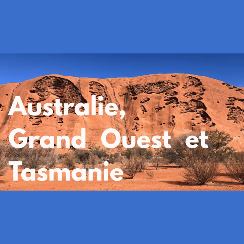 A vast, reddish rock formation dominates the background, with sparse, dry brush in the foreground under a clear blue sky.