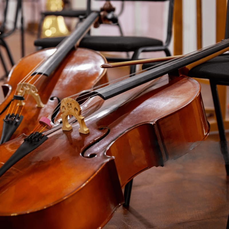 Two cellos rest on a wooden floor in front of empty chairs, with a bow lying across the foreground instrument.
