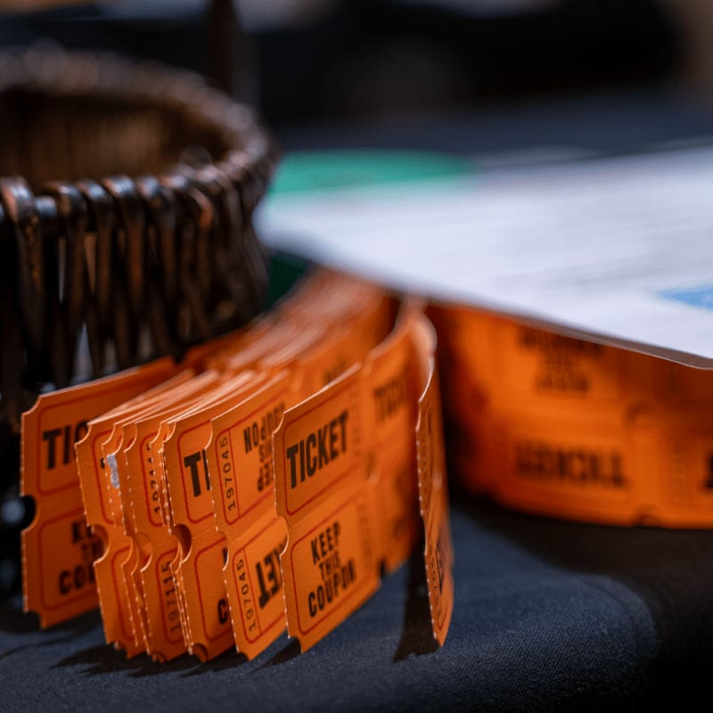 A stack of orange tickets lies in the foreground next to a woven basket, with a blurred background of papers and a green surface.
