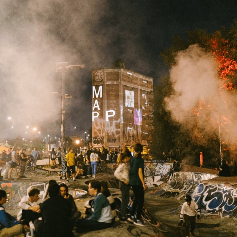 A group of people gather in a graffiti-covered skate park at night, with a tall building illuminated by projections in the background.