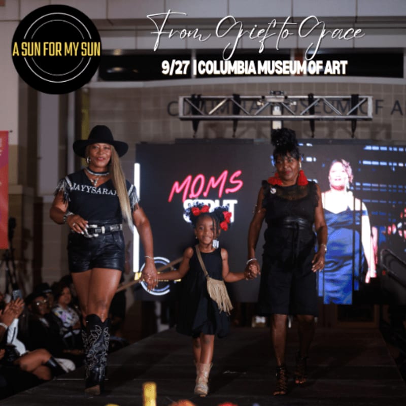 Three women, two adults and a child, walk down a runway in front of a large screen displaying "MOMS" and event details.