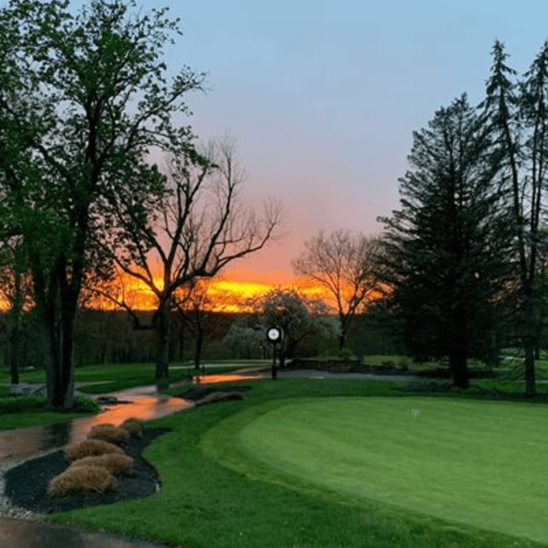 A vibrant sunset paints the sky behind a lush green golf course with a wet pathway in the foreground.