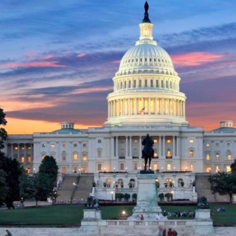 A statue stands in the foreground before the illuminated United States Capitol building, set against a vibrant sunset sky.