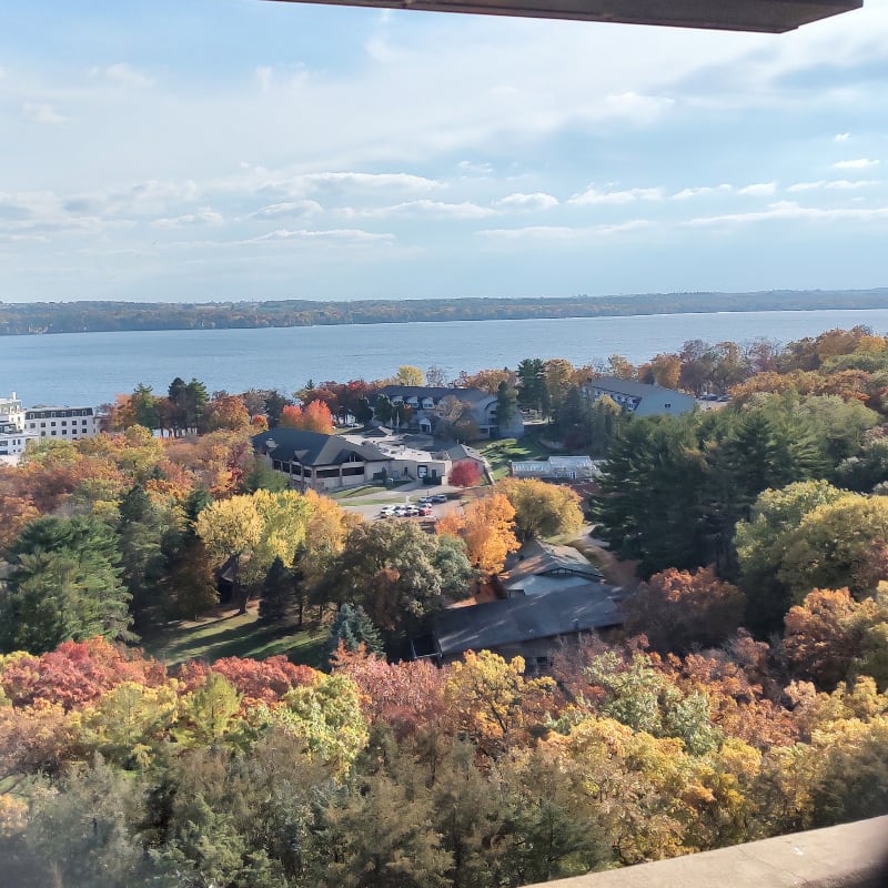 Vibrant autumn foliage surrounds buildings nestled beside a calm lake under a partly cloudy sky.