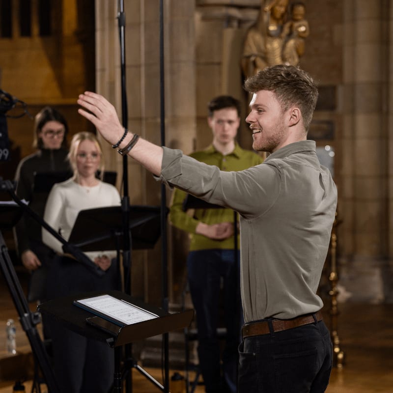 A choir director gestures with his right hand while a group of singers stand behind him in a dimly lit church.