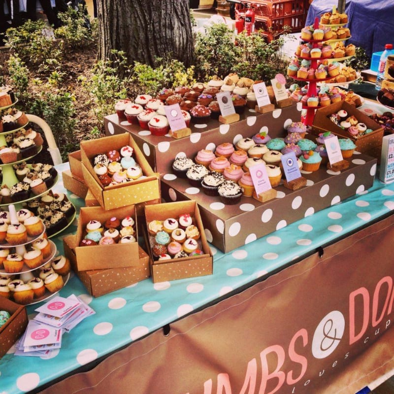 A variety of colorful cupcakes are displayed on tiered stands and in boxes on a table covered with a polka-dot cloth, with trees and more food stalls visible in the background.