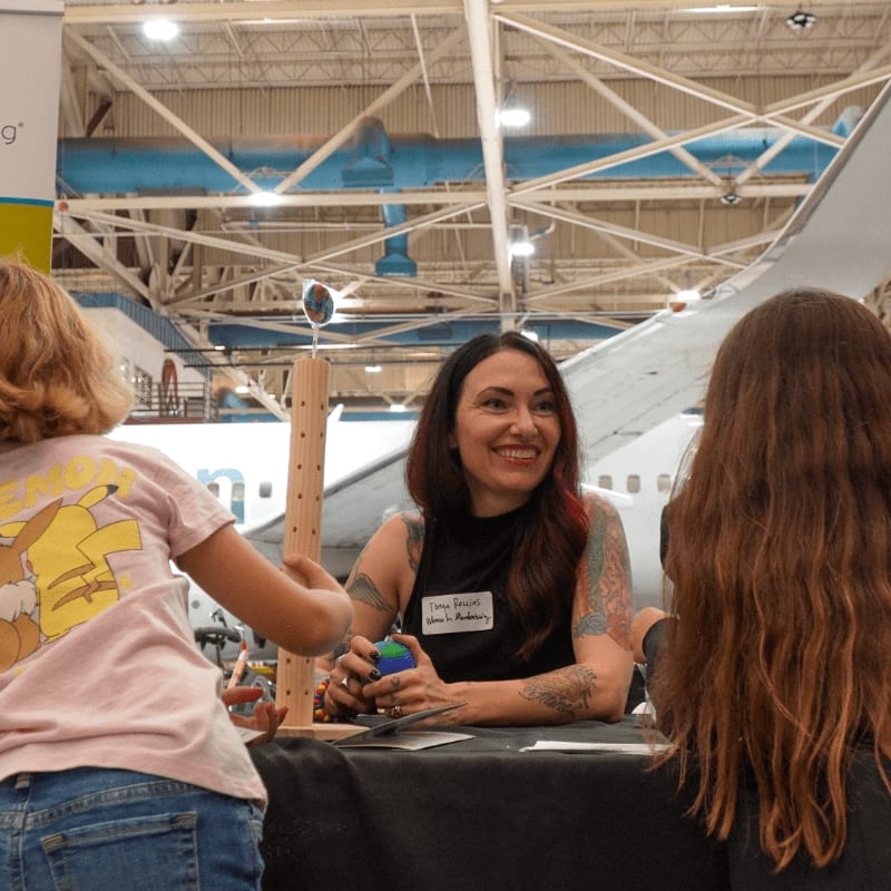 A woman with tattoos smiles at two children in the foreground, while a large airplane wing is visible in the background.