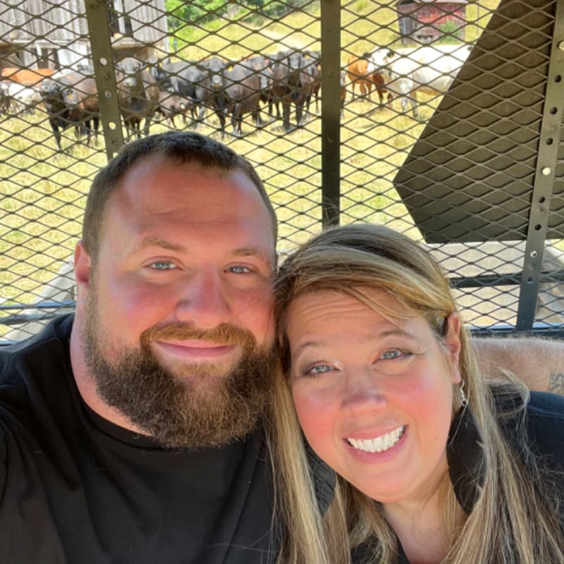 A smiling couple is in the foreground, with a herd of cattle visible behind a fence in the background.