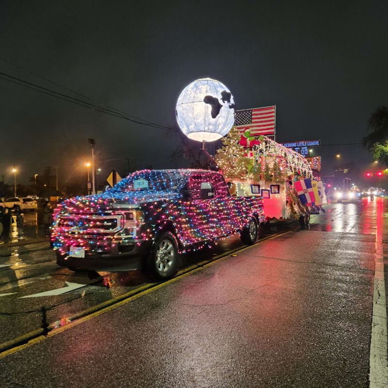 A pickup truck and trailer, both adorned with colorful Christmas lights, are parked on a wet street at night, with a large illuminated globe and an American flag visible in the background.
