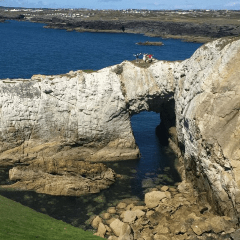 A natural rock arch frames a clear blue sea with a few people standing on top, while rugged cliffs and distant buildings form the background.
