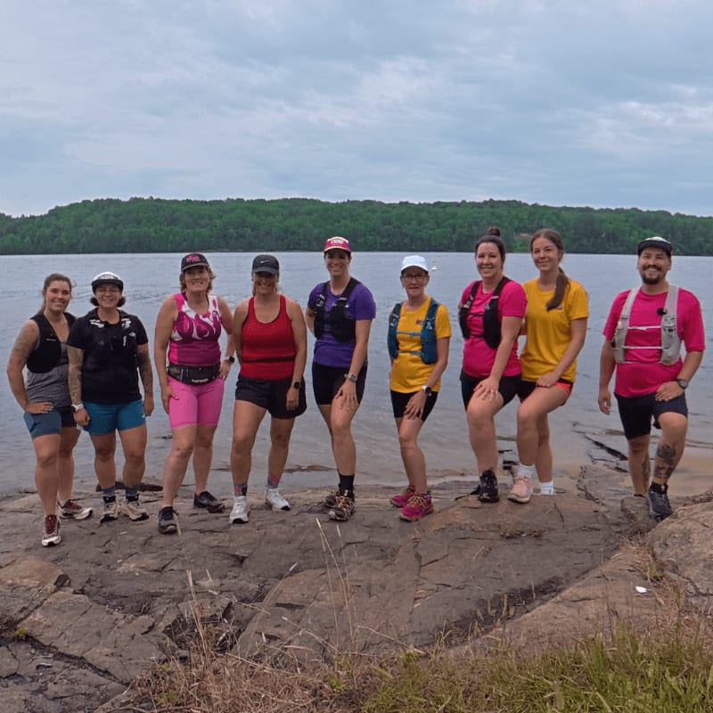 A group of nine runners in athletic wear stand on a rocky shore with a calm lake and tree-lined horizon behind them.