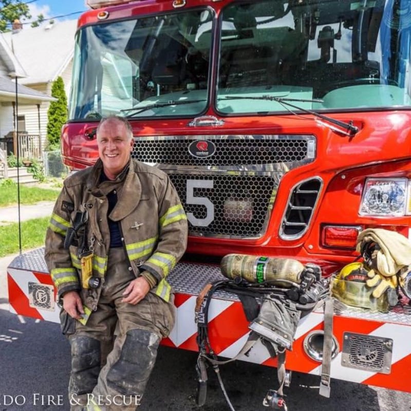 A smiling firefighter in full gear sits on the front bumper of a red fire truck, with a residential street visible in the background.