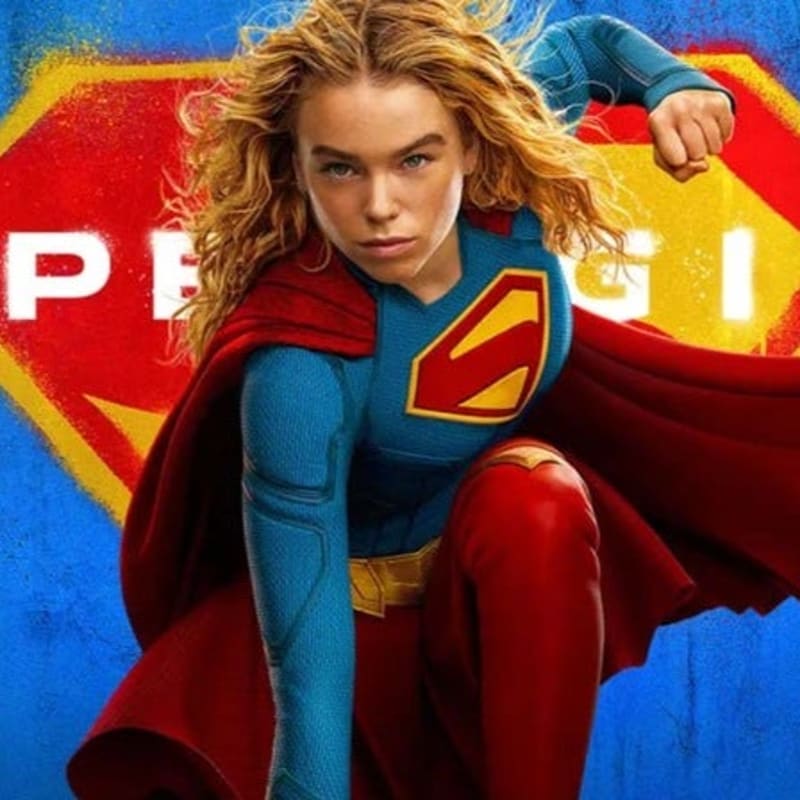 A young woman in a Supergirl costume with a red cape and blue suit is crouched forward against a backdrop of a yellow and red "S" shield.