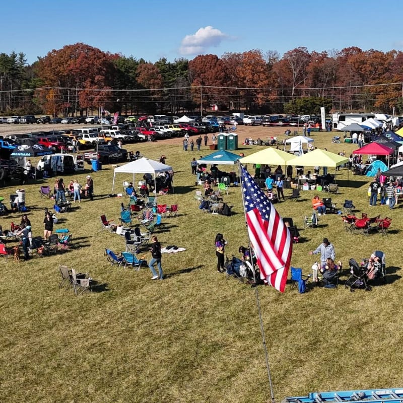 An American flag waves in the foreground of a grassy field filled with people relaxing in chairs and under tents, while a large crowd and many vehicles are visible in the background with autumn trees lining the horizon.
