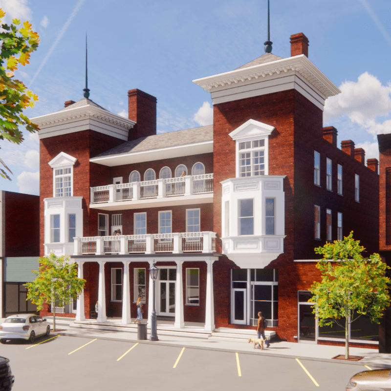 A red brick building with white trim and balconies stands on a street with a parked car and trees in the foreground, under a bright blue sky with scattered clouds.