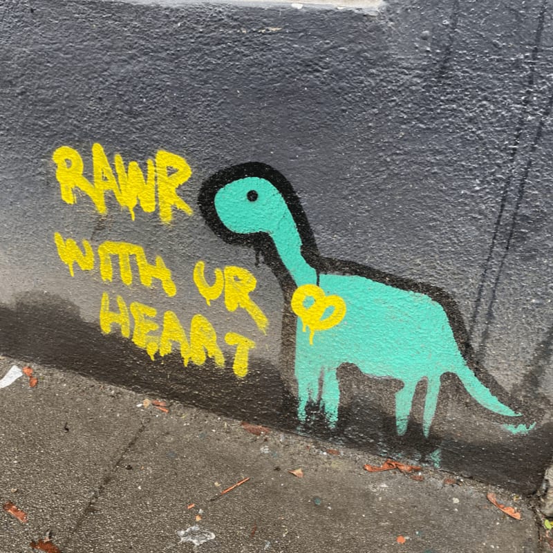 A teal dinosaur with a yellow pretzel around its neck is spray-painted on a dark gray wall, with the words "RAWR WITH UR HEART" in yellow spray paint to its left, all set against a concrete sidewalk in the foreground.
