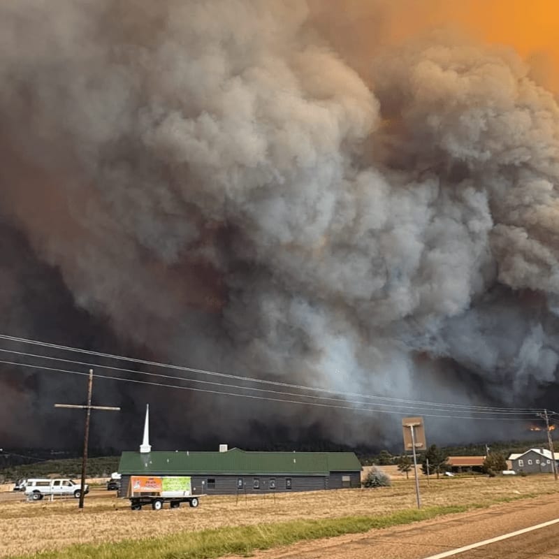 A massive plume of dark smoke billows into the orange sky behind a small town with a church and buildings in the foreground.