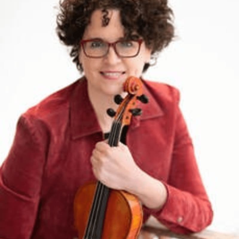 A woman with curly hair and glasses in a red jacket holds a violin in front of a blurred white background.