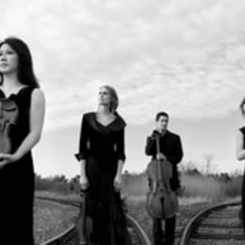 Four musicians in formal attire stand on railroad tracks with their instruments, against a cloudy sky.