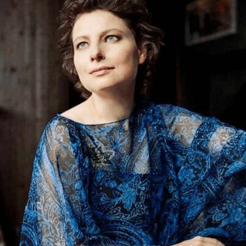 A woman with short brown hair and blue eyes, wearing a blue patterned top, looks upwards with a blurred, dark background.