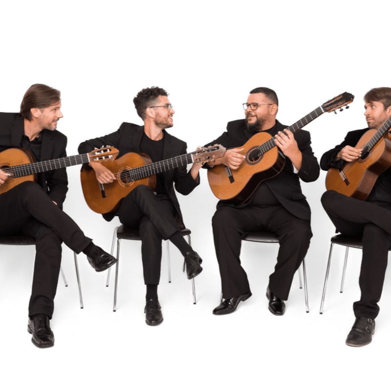 Four men in black suits sit on chairs playing acoustic guitars against a plain white background.