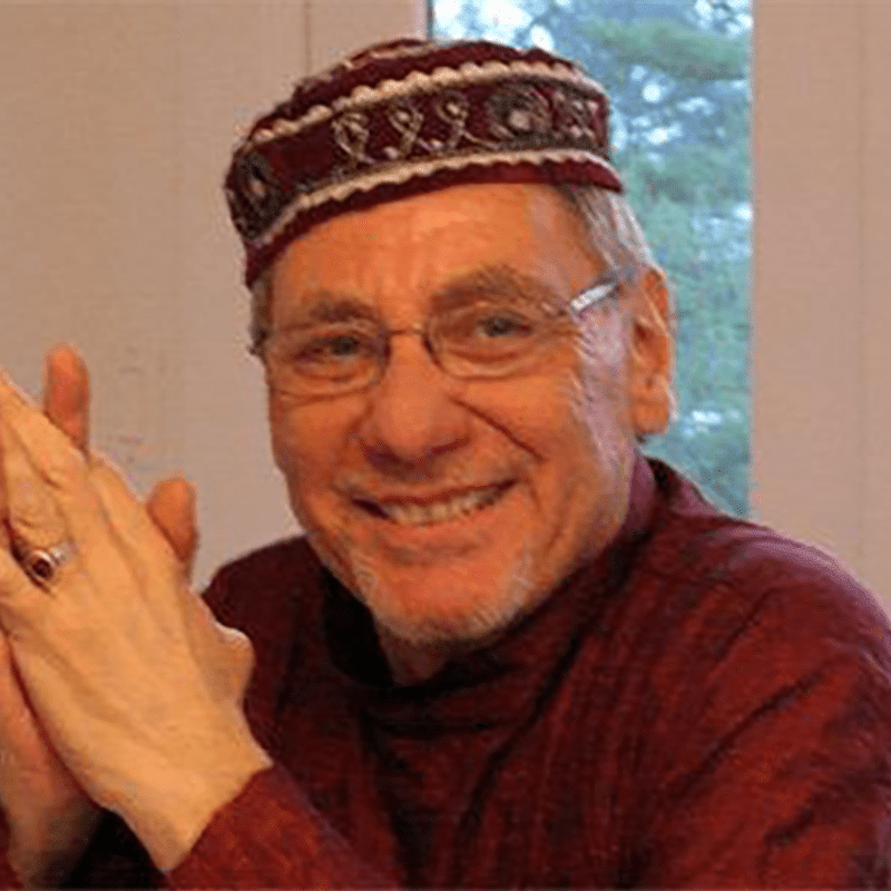 A smiling man in a maroon cap and shirt is in the foreground, with a blurred window and trees in the background.