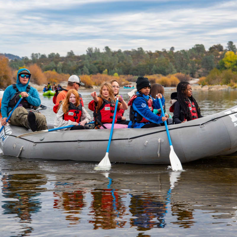 A group of children and adults in a raft paddle down a calm river with trees and hills in the background.