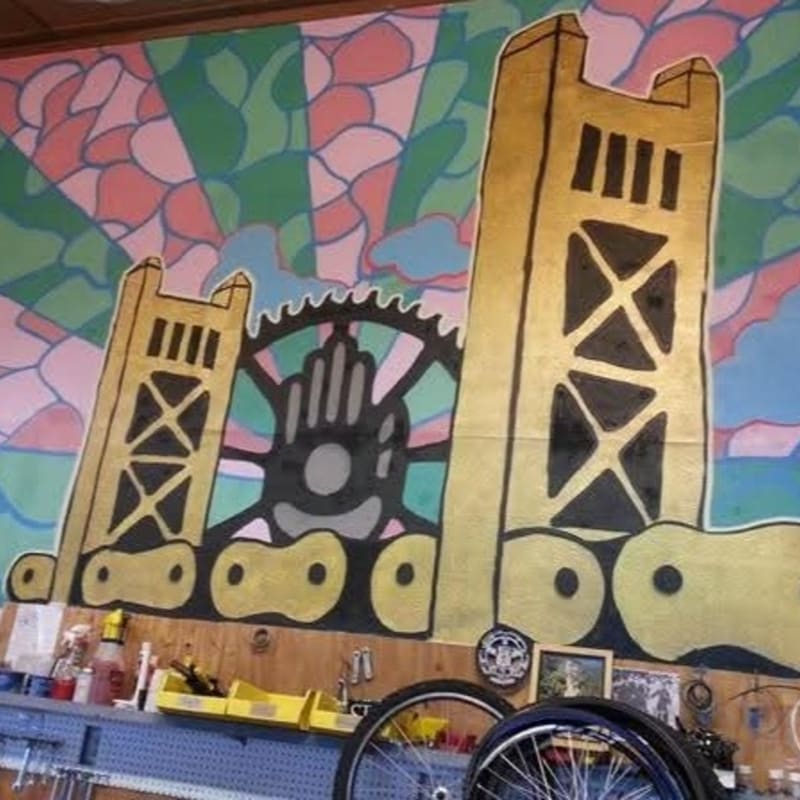 A vibrant mural of a golden bridge and bicycle gears adorns the wall above a workbench cluttered with tools and bicycle parts.