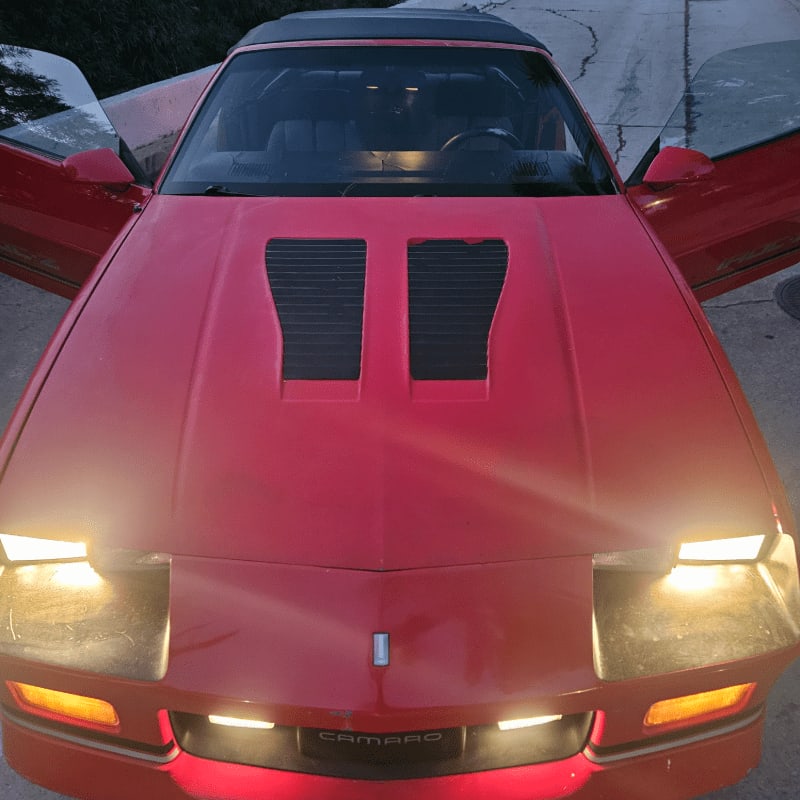 A red Camaro with its doors and headlights open sits on a paved surface with a dimly lit background.
