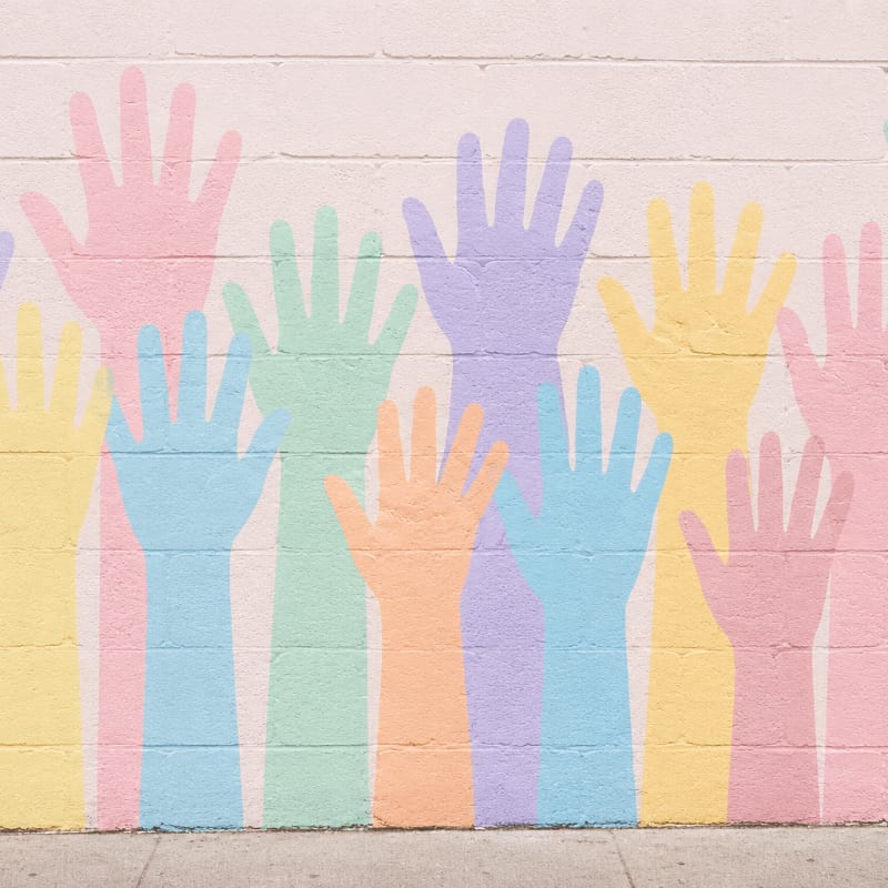 Pastel-colored hand silhouettes are painted on a white brick wall above a sidewalk.