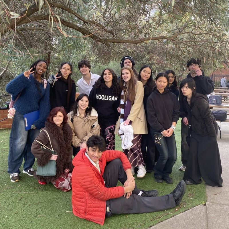 A group of teenagers poses for a photo on a grassy area with trees and a building in the background.