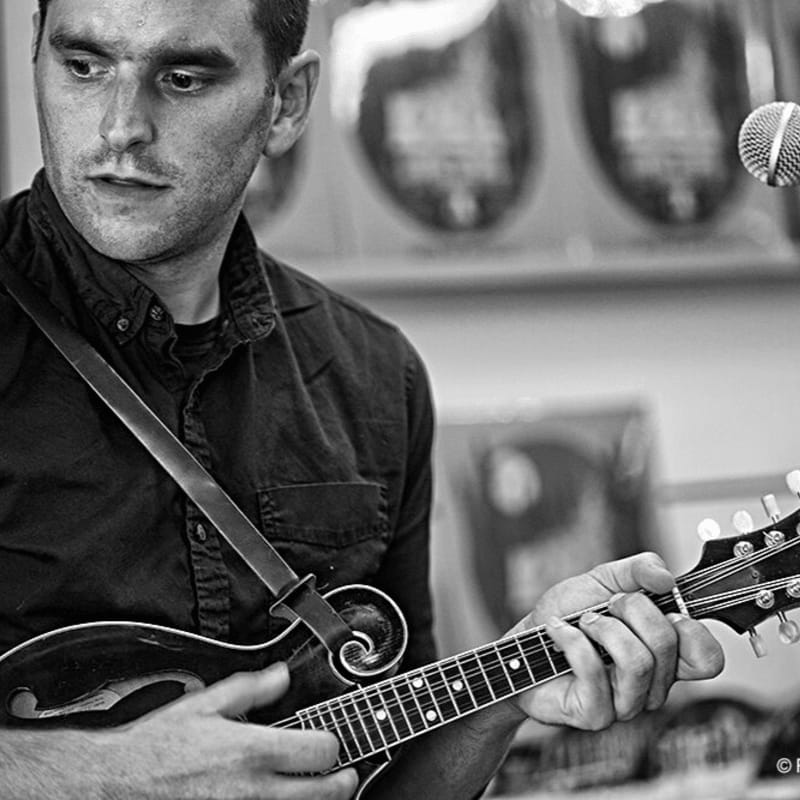 A man plays a mandolin in the foreground, with a microphone and blurred records in the background.