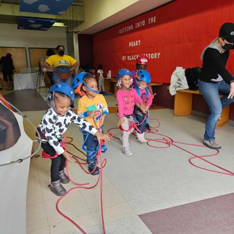 Young children in helmets and harnesses stand in the foreground with red ropes, while a red banner reading "GETTING INTO THE HEART OF BLACK HISTORY" hangs in the background.